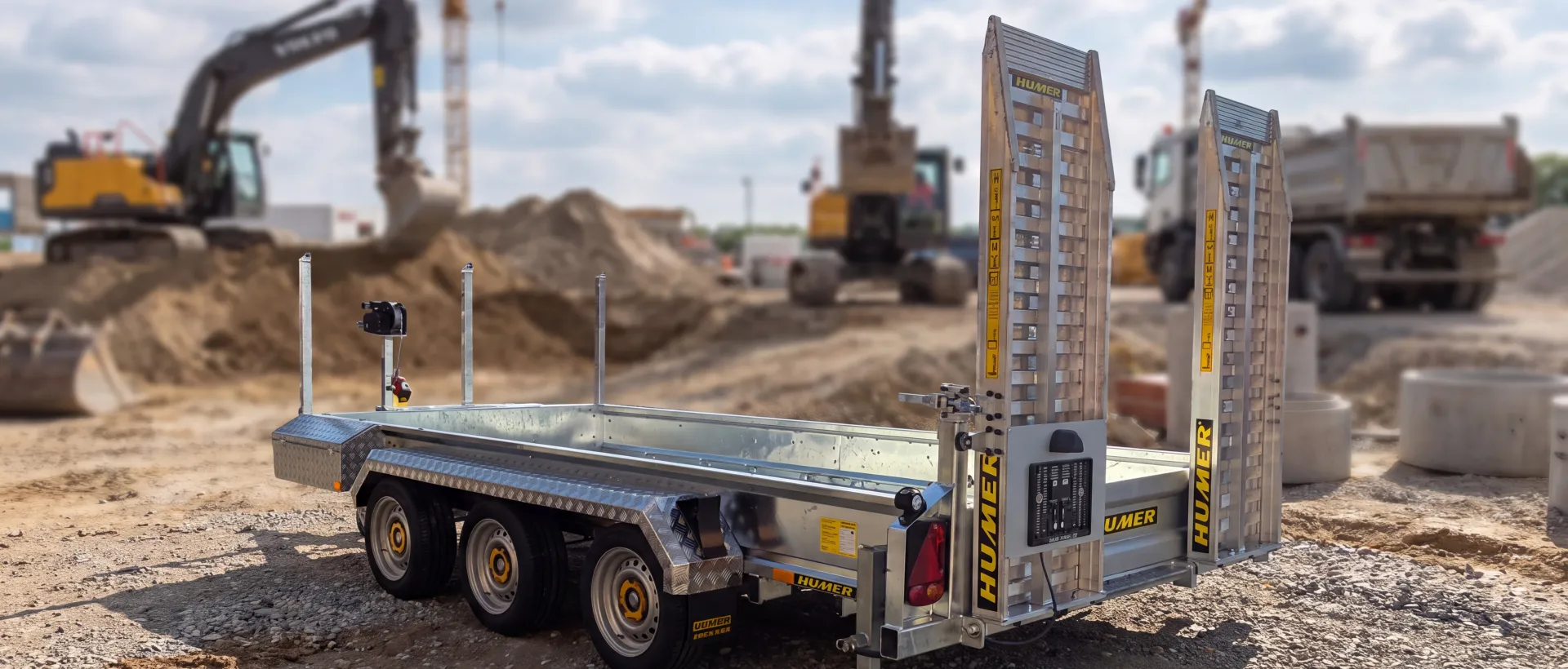 Humer Tieflader-Baumaschinenanhänger mit drei Achsen und aufrechten Auffahrrampen auf einer Baustelle mit Baggern und Lkw im Hintergrund.