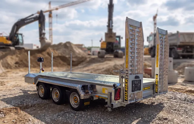 Humer Tieflader-Baumaschinenanhänger mit drei Achsen und aufrechten Auffahrrampen auf einer Baustelle mit Baggern und Lkw im Hintergrund.