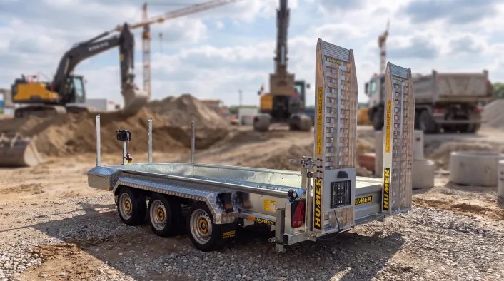 Humer Tieflader-Baumaschinenanhänger mit drei Achsen und aufrechten Auffahrrampen auf einer Baustelle mit Baggern und Lkw im Hintergrund.